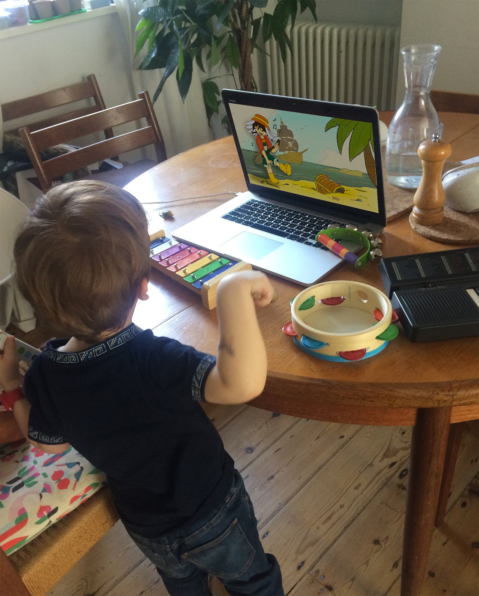 A young child stands at a table playing a xylophone and tambourine while watching a Daisy Digs It animation on a laptop